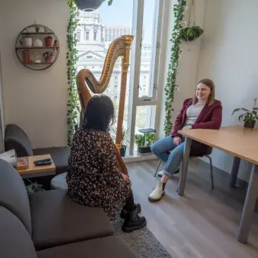 two students talk in their dorm, one sits in front of a harp