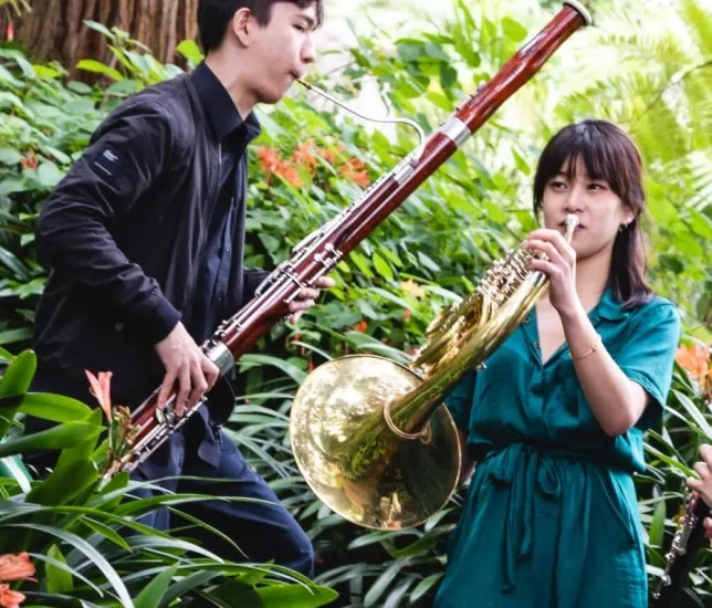 students play a bassoon and a horn among many green plants