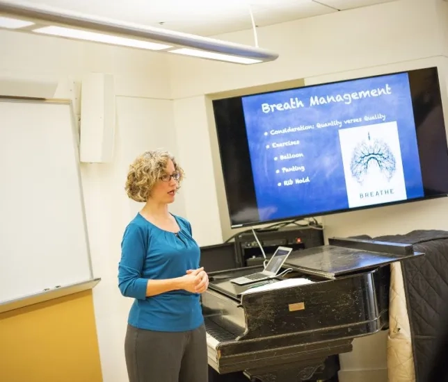 a professor lectures about vocal health with an anatomy chart behind her