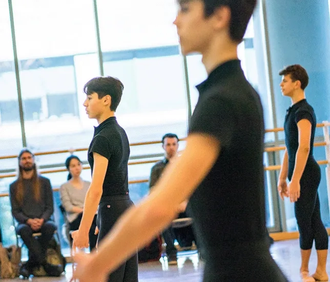 Three ballet dancers wearing black clothes perform in a studio space