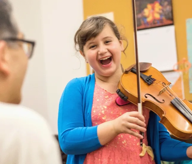 A young girl smiles while practicing violin with her teacher.