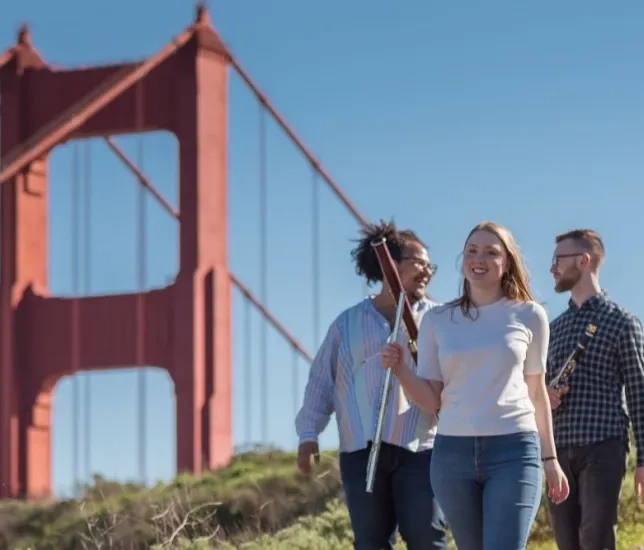 students hold their instruments and walk along a trail where the tops of the golden gate bridge is visible behind them