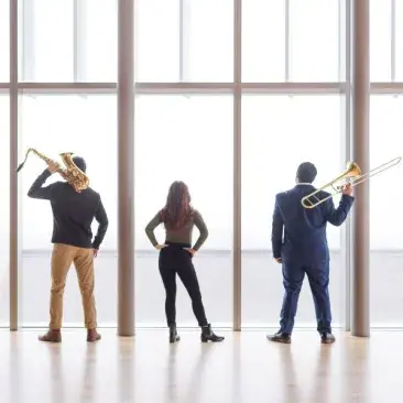 students stand with their backs to the camera, holding their instruments, looking out the 11th floor of bowes window