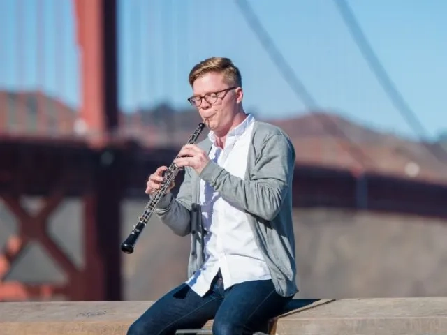 an oboe player sits on a ledge while playing the oboe in front of the golden gate bridge