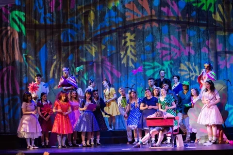 a performer is slouched over a desk while many ensemble members sing around them