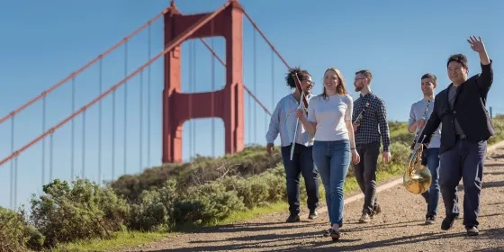A group of students with instruments walk down a path with the Golden Gate Bridge visible in the background.
