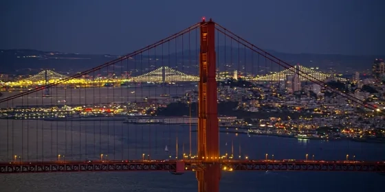 Aerial night view of the Golden Gate Bridge's north tower, with the illuminated San Francisco skyline, Bay Bridge, and bay   waters in the background.