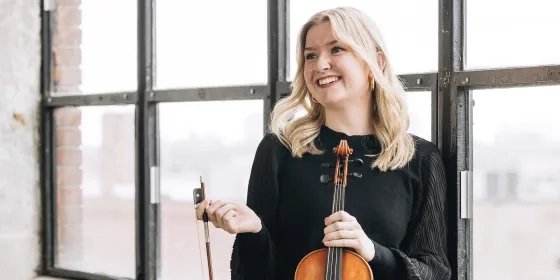 A smiling SFCM student holds a violin in front of a window.