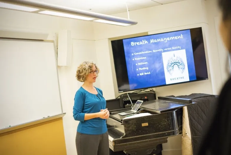 a professor lectures about vocal health with an anatomy chart behind her