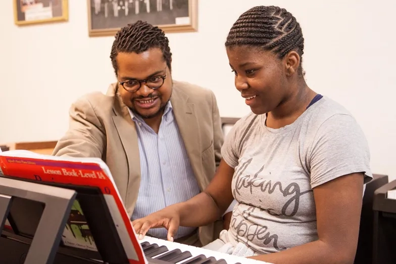 A male teacher with a female student at a keyboard with the teacher pointing to some sheet music